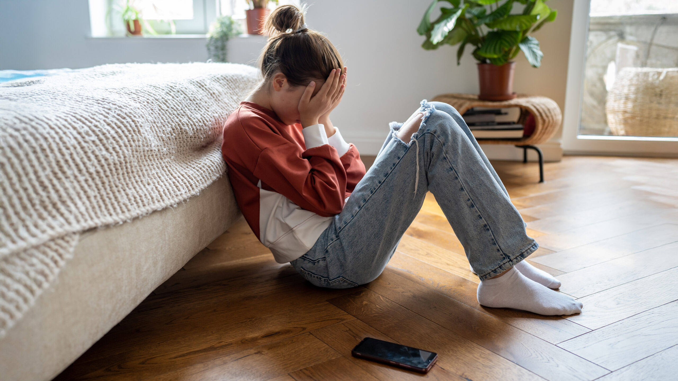 15-årsgrense for sosiale medier: Er det allerede for sent? 1 Unhappy teen girl covering face with hands crying while sitting on floor with mobile phone nearby