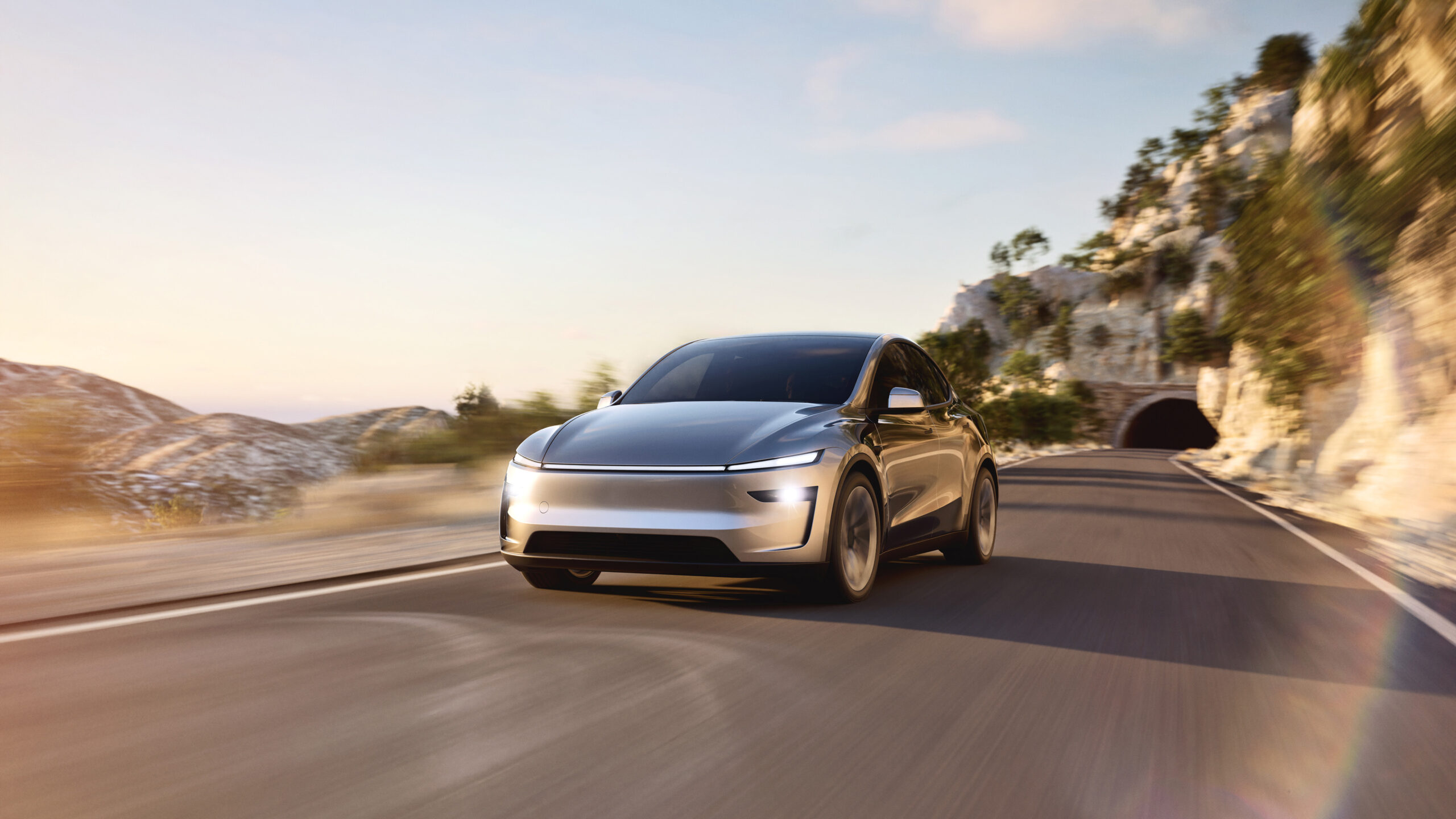 Filholderassistenten i Tesla krever snart abonnement 2 Sølvfarget Tesla Model Y kjører på en svingete asfaltvei langs en fjellside med en tunnelåpning i bakgrunnen.