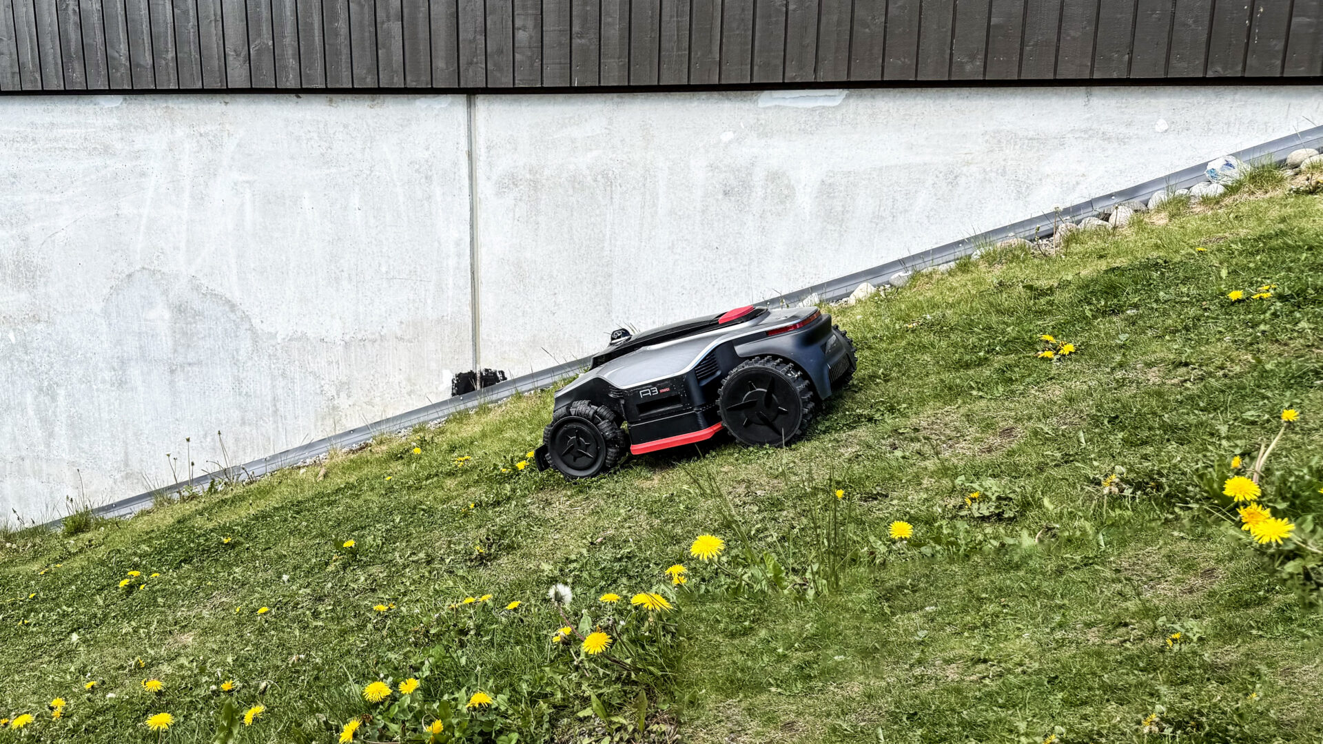 Dreame A3 AWD Pro 2500 11 Dreame A3 AWD Pro klatrer rolig nedover en bratt gresskledd skråning, like ved en grålig betongvegg med svart husfasade over.