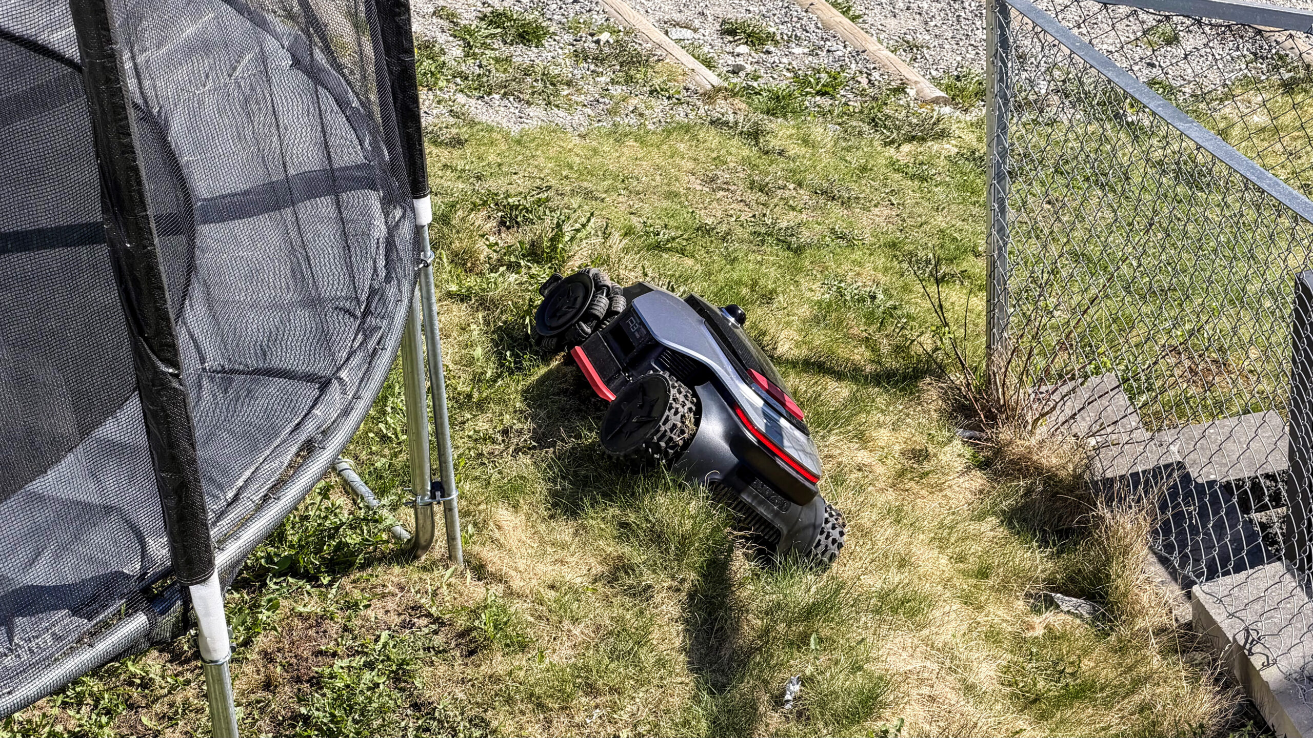 Dreame A3 AWD Pro har veltet på siden i en smal stripe gress mellom en trampoline og et flettverksgjerde, etter å ha rygget for nært en oppbygd kant.