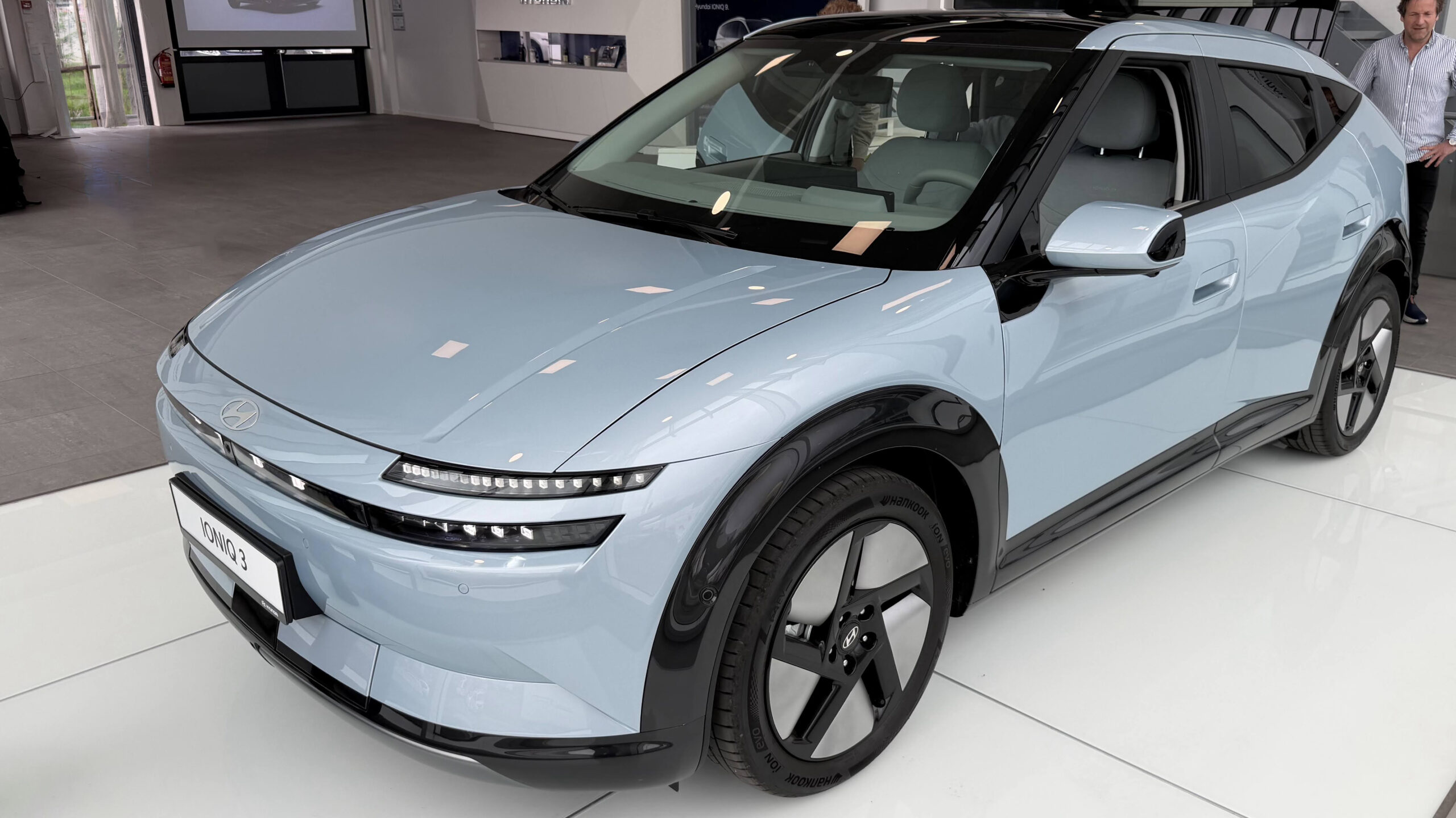 Ioniq 3: Urban elbil med smart teknologi 1 Lyseblå Hyundai Ioniq 3 fotografert skrått forfra innendørs, med den horisontale LED-lyslisten og pikselgrafikken i fronten godt synlig.