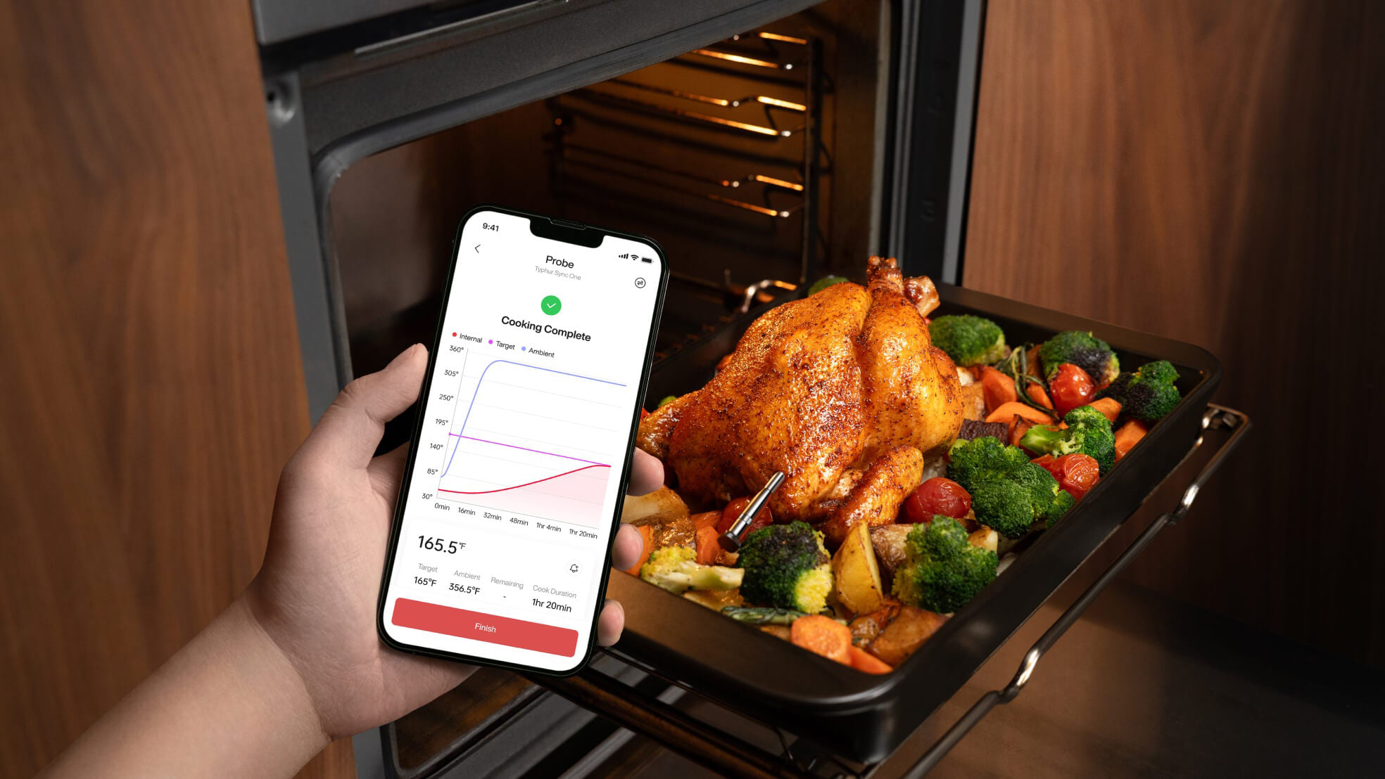 Typhur Sync Gold steketermometer: Alle får biffen som de vil 3 En hånd holder en smarttelefon foran en åpen stekeovn. Skjermen viser Typhur-appen med en temperaturkurve, måltemperatur 165 grader Fahrenheit og meldingen "Cooking Complete". I ovnen står en hel stekt kylling med brokkoli, poteter, gulrøtter og tomater, med en sonde stukket inn i bryststykket.