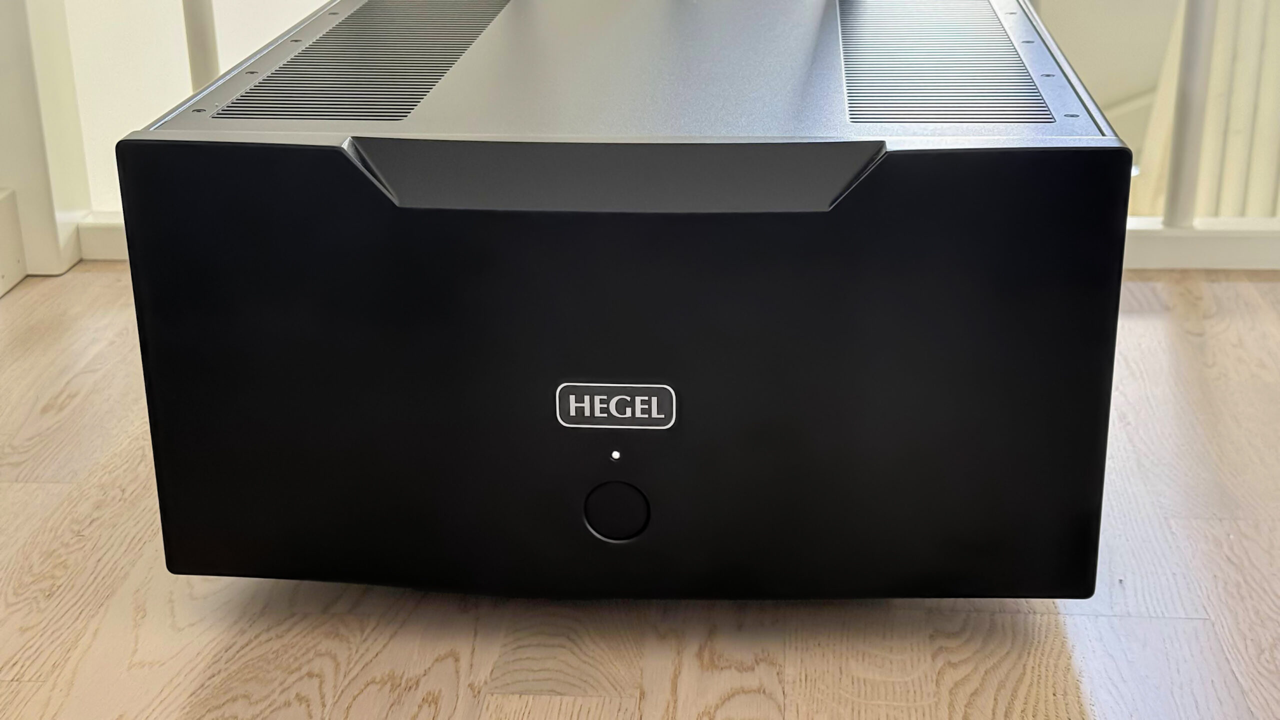 IsoTek V5 Sirius 3 Stor svart Hegel H30A effektforsterker med Hegel-logo og én rund knapp på fronten, plassert på et lyst tregulv.