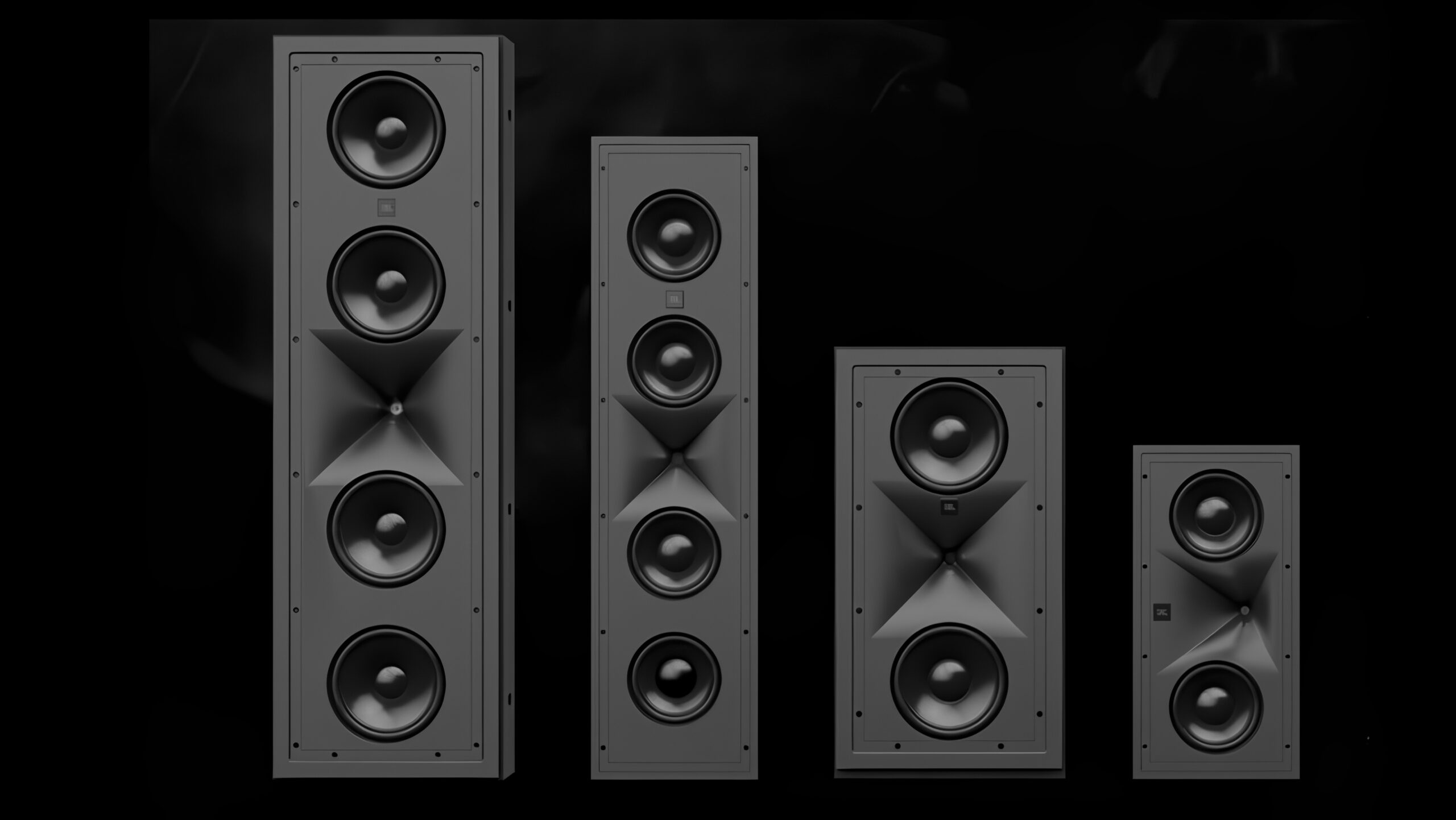 Ny JBL Synthesis: kraften du ikke ser 3 Fire grå JBL-høyttalere av ulik størrelse stilt opp på rad mot sort bakgrunn, fra en høy modell med fire basselementer til venstre til en kompakt modell til høyre. Alle har synlige basskjegler og en hornformet diskant uten frontgitter.