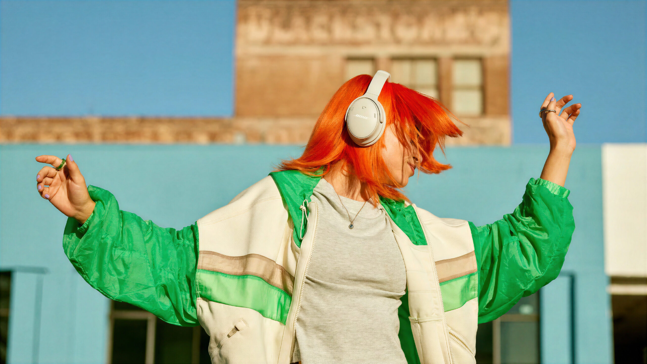 Lekkasje: Nye Bose QuietComfort kommer om få uker 1 Kvinne med oransje hår danser utendørs med hvite Bose QuietComfort Headphones (2nd Gen) på hodet, kledd i grønn og hvit jakke foran en murbygning.